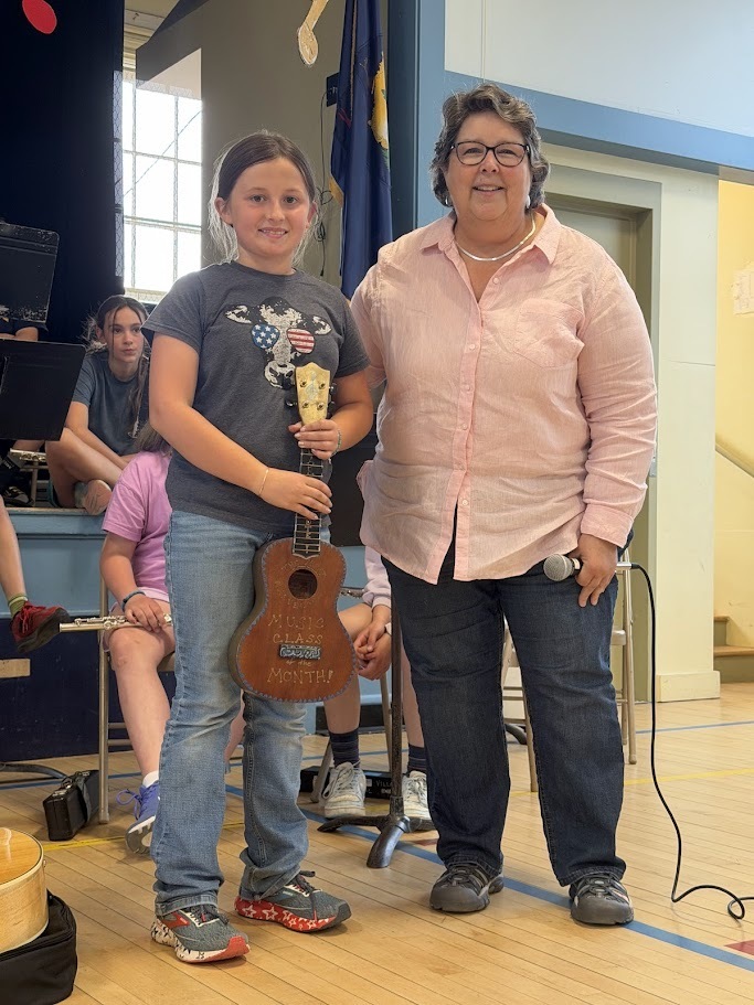 A student stands beside a teacher during an awards presentation at an Orwell Village School music assembly. The student holds a decorated ukulele-style instrument while smiling for a photo. Other students and musical instruments are visible in the background.