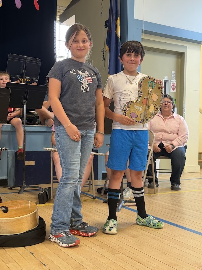Two elementary students stand together during an awards presentation at an Orwell Village School assembly. One student holds a colorful, decorated guitar-shaped art project while classmates and music stands are visible behind them.