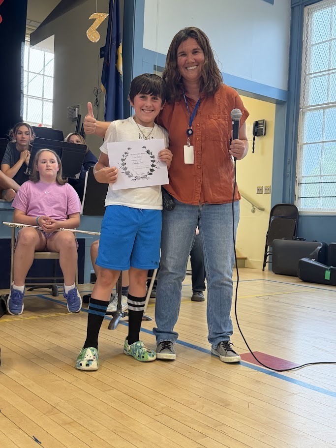 A student gives a thumbs-up while standing next to a staff member and holding a certificate during an Orwell Village School assembly. Seated students and music stands are visible behind them.