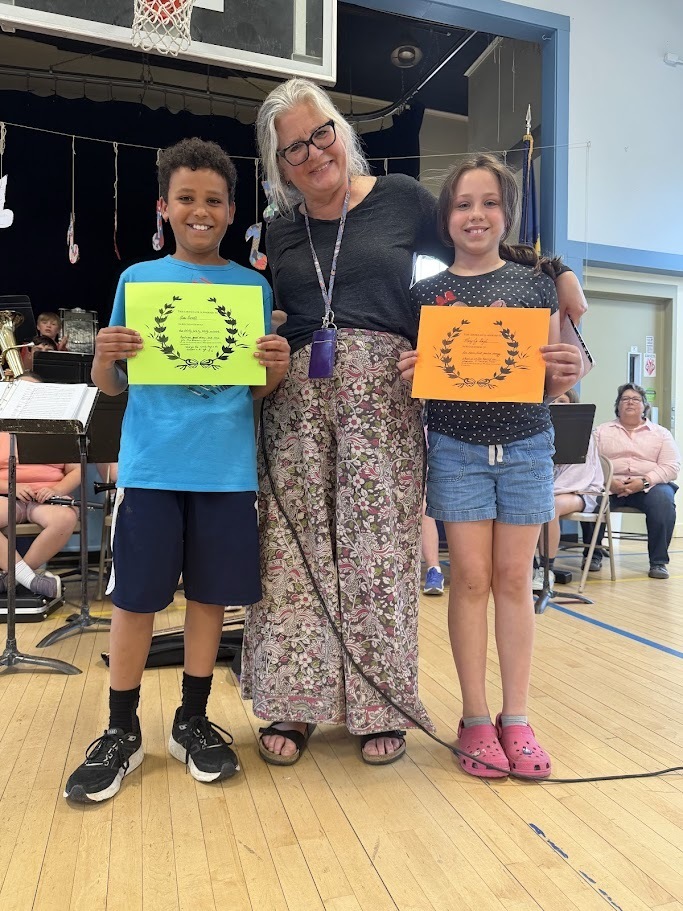 A staff member poses with two students, each holding a colorful certificate, during an Orwell Village School awards ceremony. The group smiles while standing in front of the school band.