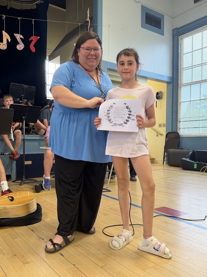 A student stands with a staff member while holding a certificate during an awards presentation at Orwell Village School. The pair smile for a photo in front of the assembled band.