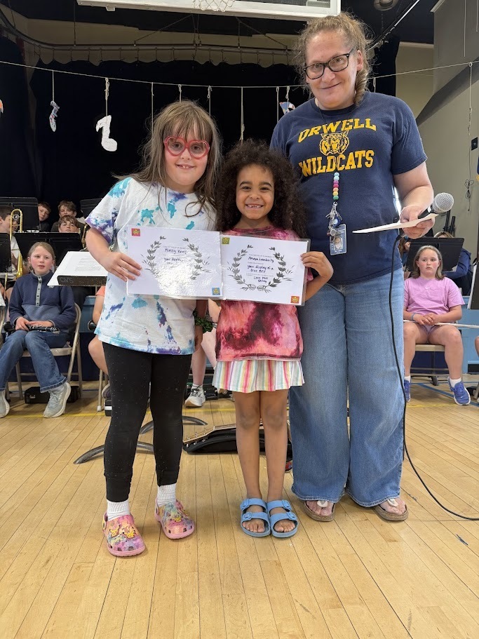 Two elementary students stand with a staff member while holding certificates during an Orwell Village School awards assembly. Students from the band are seated in the background.