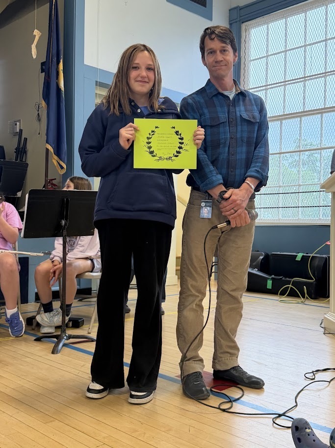 A student holds a bright green certificate while standing beside a staff member during an Orwell Village School awards presentation. The pair pose for a photo in the school gymnasium with band equipment visible nearby.