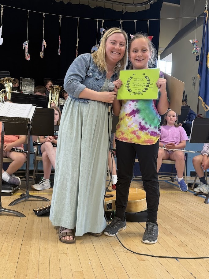 A staff member stands beside a student holding a bright green certificate during an Orwell Village School assembly. Musical instruments and student performers are visible in the background.