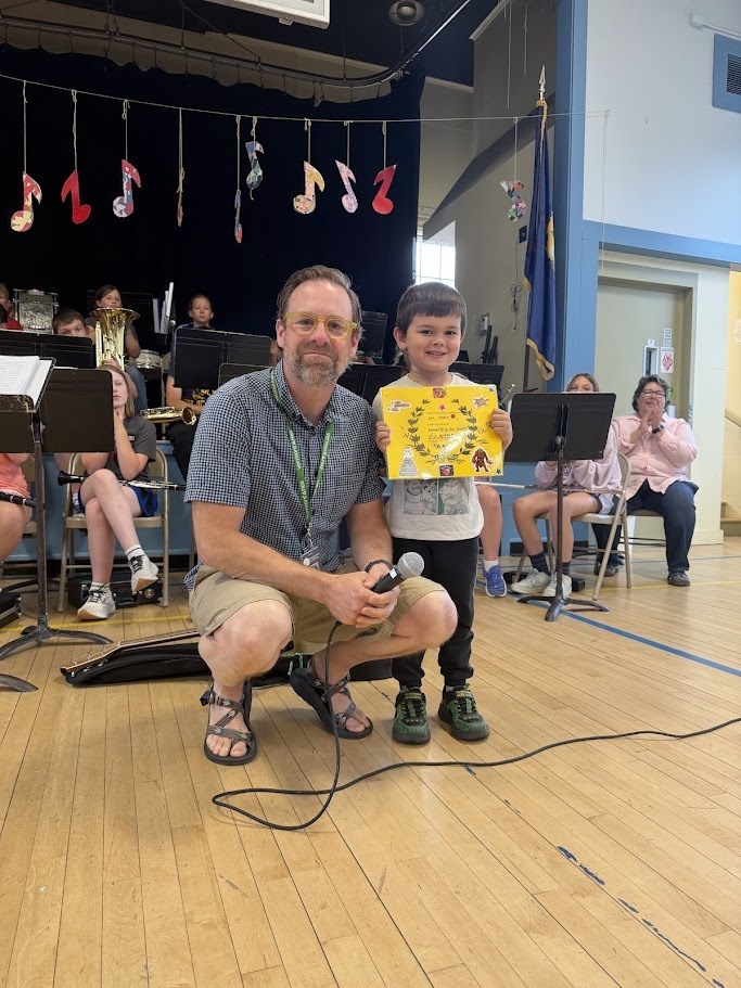 An adult kneels beside a smiling student holding a colorful certificate during an Orwell Village School assembly. Members of the school band sit behind them with instruments and music stands.
