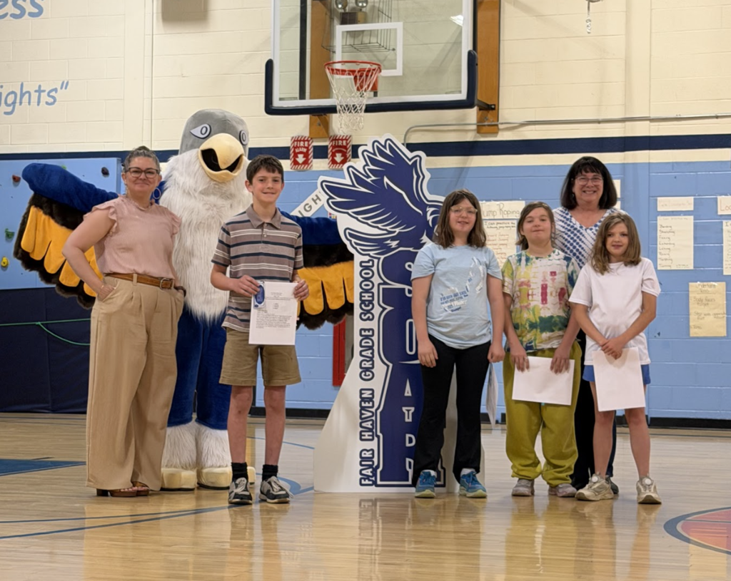 A group of students and staff members pose in the Fair Haven Grade School gymnasium beside the school mascot and a large Falcon display during a student recognition ceremony. Several students hold certificates.