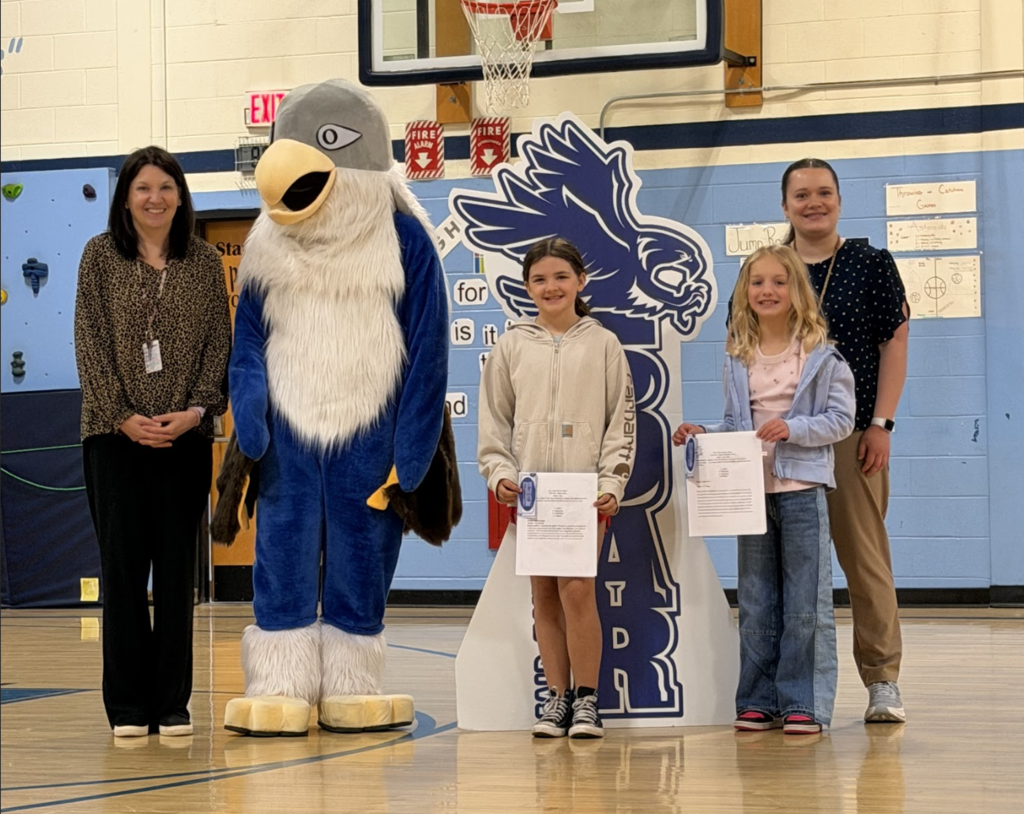 Two students holding certificates stand beside two staff members and the Falcon mascot during an awards ceremony in the school gym.