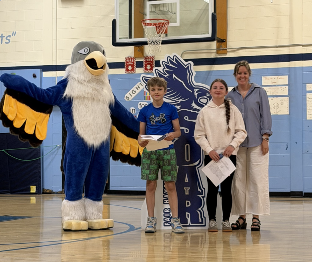 Two students stand with a staff member and the Falcon mascot in the school gymnasium while holding recognition papers during an awards assembly.
