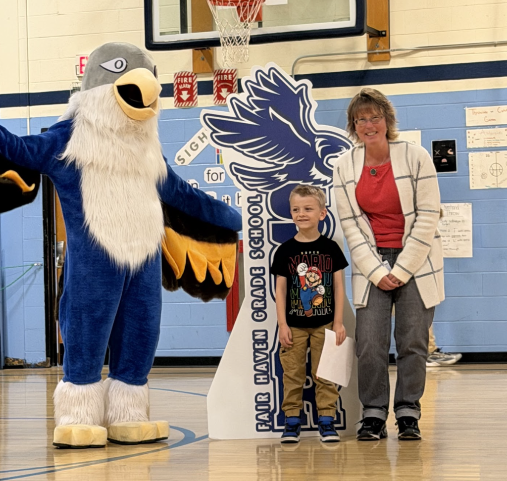 A young student stands with an adult and the Falcon mascot beside a large Falcon display during a school awards presentation.