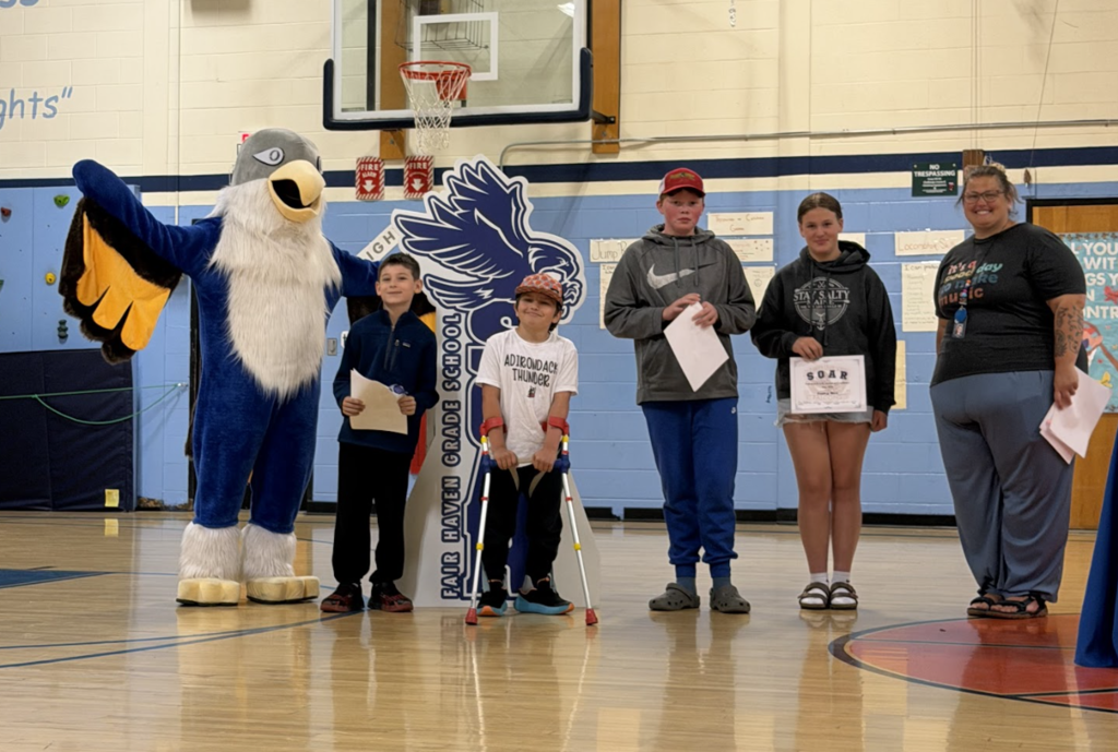 Four students, including one using forearm crutches, stand with a staff member and the Falcon mascot during a school recognition ceremony.