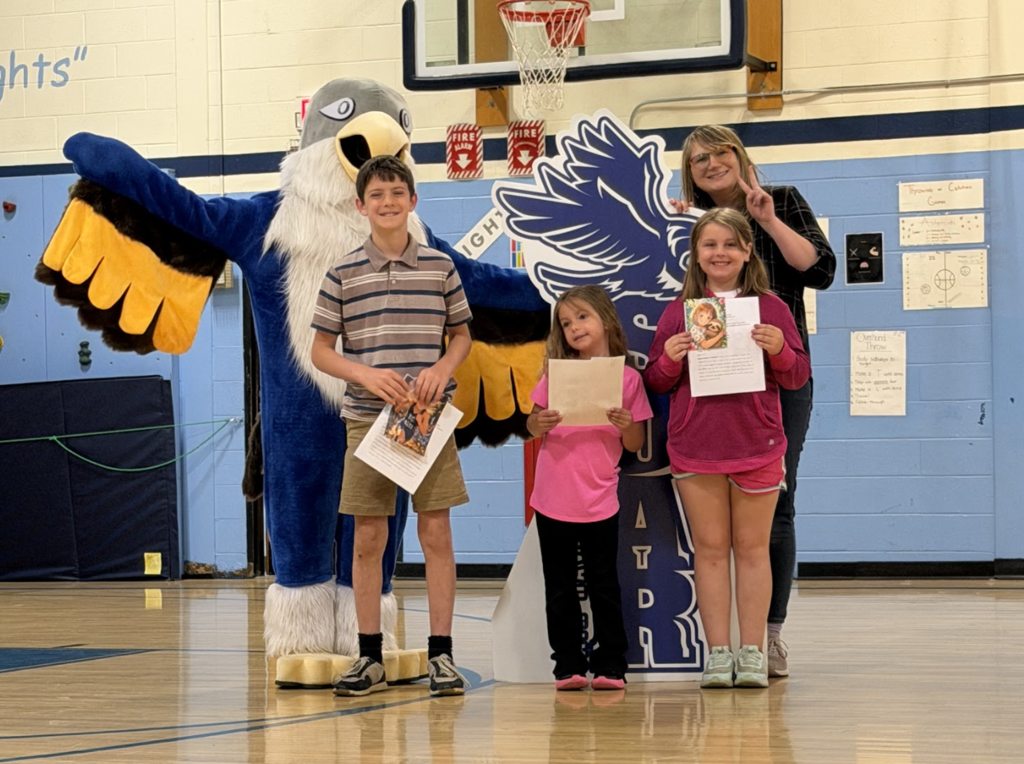 Three students holding papers pose with a staff member and the Falcon mascot in the gymnasium during a student awards assembly.