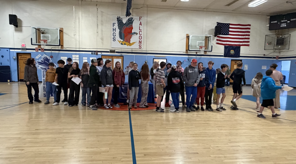 A large group of middle school students gathers in the Fair Haven Grade School gymnasium during an assembly. Students stand in line near a table while classmates and staff move throughout the gym.