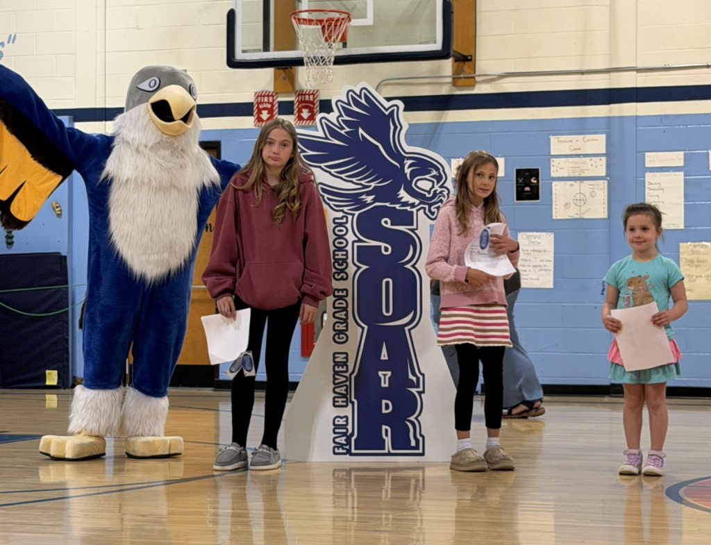 Three students stand in front of the Falcon display and mascot, holding recognition papers during a Fair Haven Grade School awards ceremony.