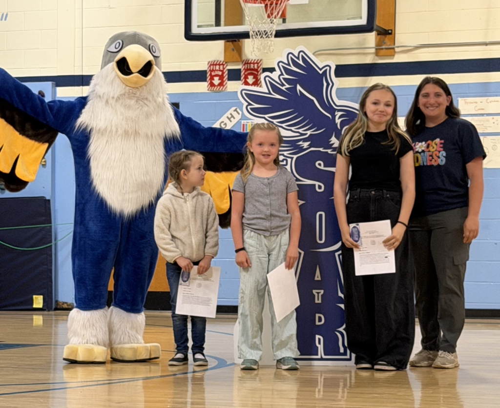 Three students of varying ages stand with a staff member and the Falcon mascot while holding certificates and recognition papers during an assembly.