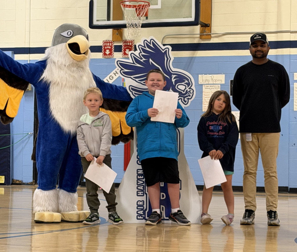 Three students holding recognition papers stand with an adult and the Falcon mascot during a Fair Haven Grade School assembly.