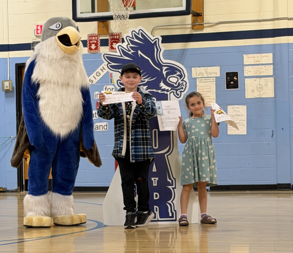Two students hold certificates and recognition papers while posing with the Falcon mascot during a student awards assembly in the gymnasium.