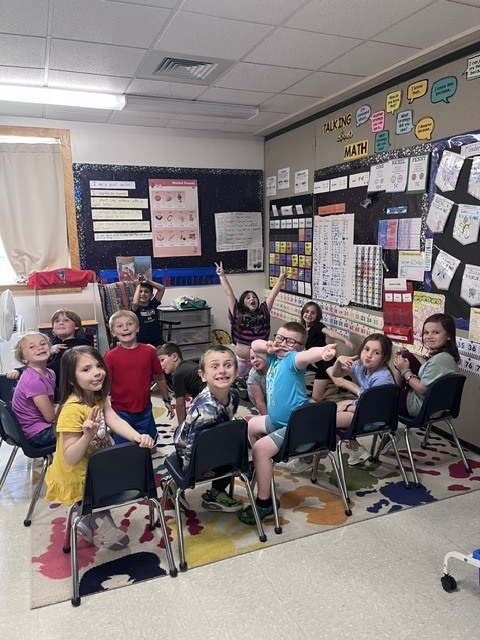 A group of about a dozen young elementary school children are gathered in a circle on a colorful, paint-splattered rug inside a classroom. Most of the kids are sitting on small navy blue chairs, while a few sit or lean on the floor. They are all looking toward the camera with energetic expressions—many are smiling widely, pulling funny faces, giving peace signs, or pointing at the camera. The classroom background features bulletin boards with educational posters about math and writing, a window with a beige curtain, and a tiled ceiling with fluorescent lighting.