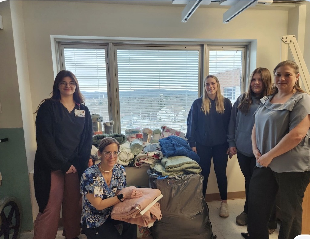 Five individuals stand around a large collection of donated blankets inside the Medical Oncology Unit at Rutland Regional Medical Center. Two Fair Haven Union High School seniors, Riley and Theresa, are pictured with hospital staff beside bags and stacks containing more than 100 donated blankets collected during the 4th Annual Blanket Drive. Large windows in the background overlook the surrounding community.