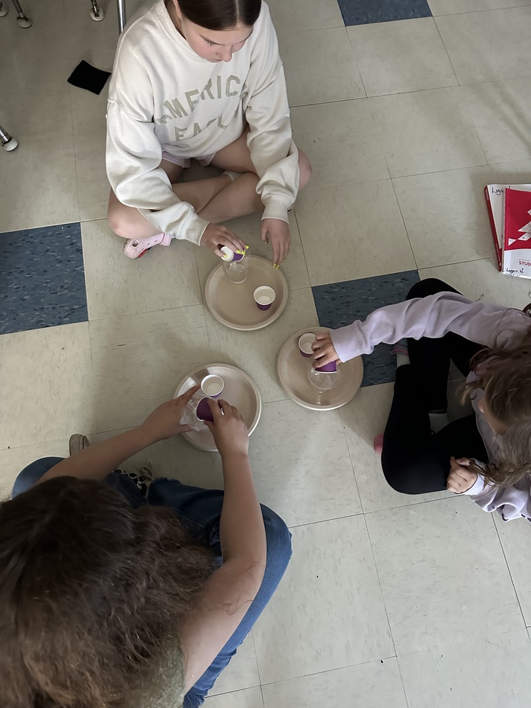 Overhead view of three fifth-grade students sitting on the classroom floor around paper plates holding cups and science materials. The students carefully pour ingredients into cups as they conduct a baking soda and vinegar experiment.
