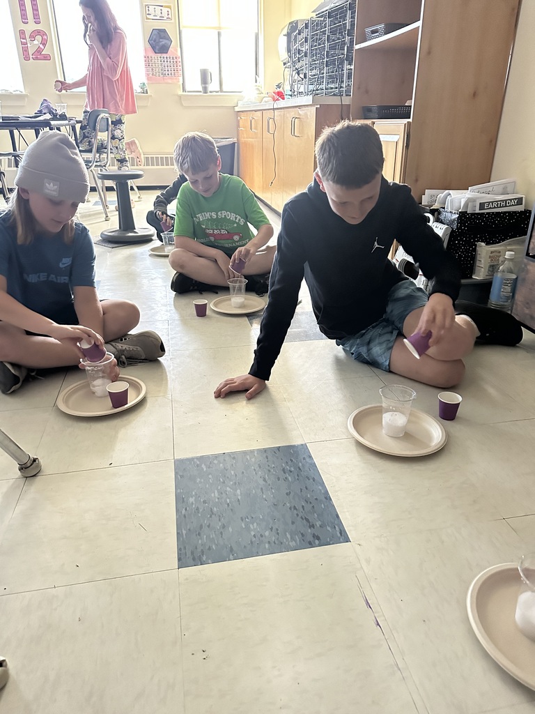 Several fifth-grade students sit on the classroom floor with paper plates and cups as they observe a bubbling chemical reaction. A teacher stands in the background while students focus on their science investigation.