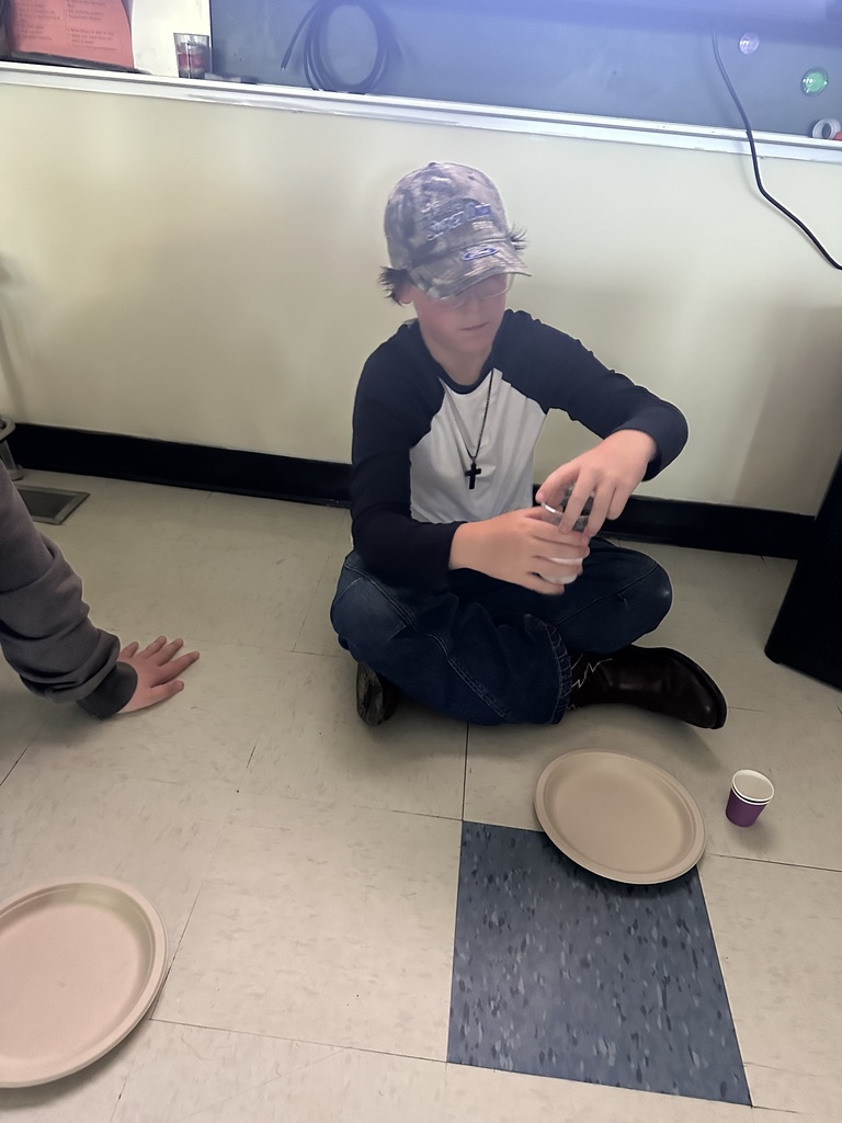 A student wearing a baseball cap sits cross-legged on the floor while holding a cup during a science investigation. A paper plate and additional materials are arranged nearby as the student participates in the baking soda and vinegar experiment.