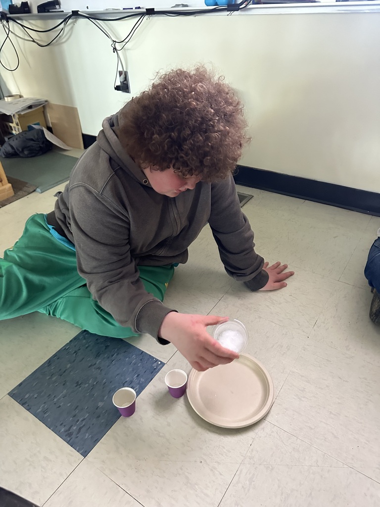 A student kneels on the classroom floor holding a cup containing a bubbling science experiment. Two small paper cups and a paper plate are nearby as the student observes the chemical reaction.