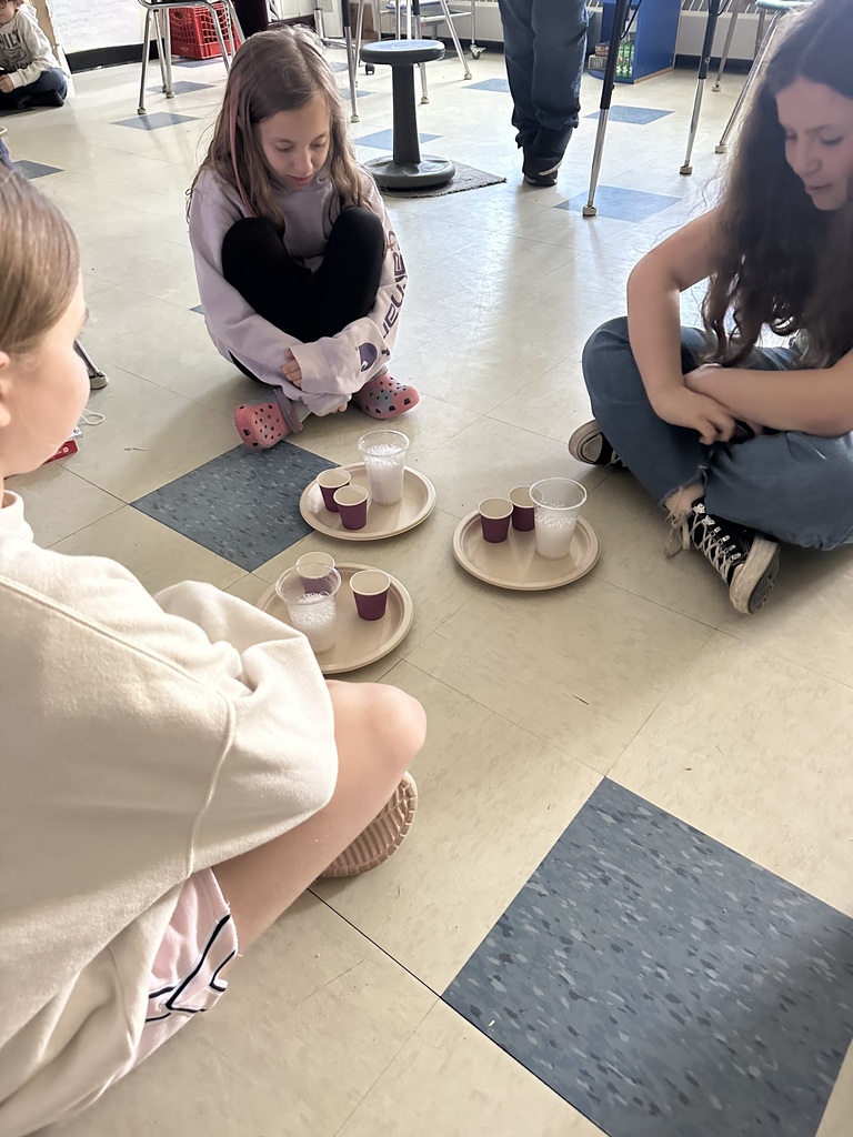 Three students sit in a circle around paper plates containing cups filled with a foamy baking soda and vinegar reaction. The students watch closely and discuss the results of their experiment.