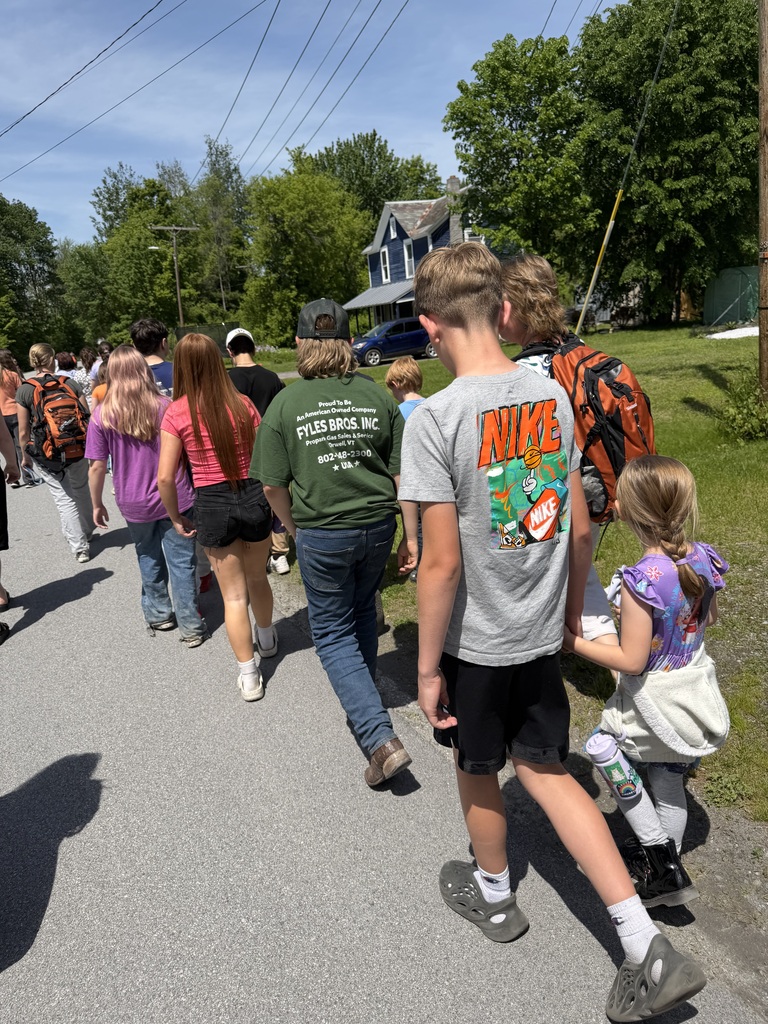 A group of Fair Haven Grade School students walks together along a roadside during a relocation drill on a sunny day. In the foreground, an older sixth-grade student walks beside a kindergarten student, offering support as they travel with classmates. Trees, homes, and utility lines can be seen in the background, highlighting the orderly and supervised nature of the drill.