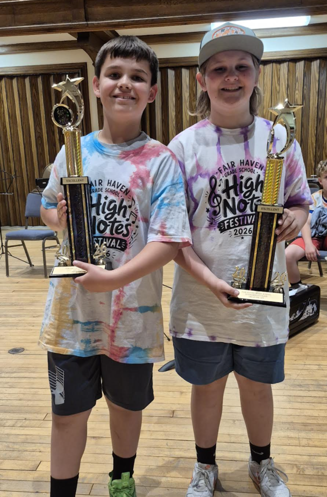 Two Fair Haven Grade School students stand on a stage holding tall trophies labeled “Excellent” during the 2026 High Notes Festival. Both students wear tie-dye festival T-shirts and smile proudly while posing with their awards in a performance hall.