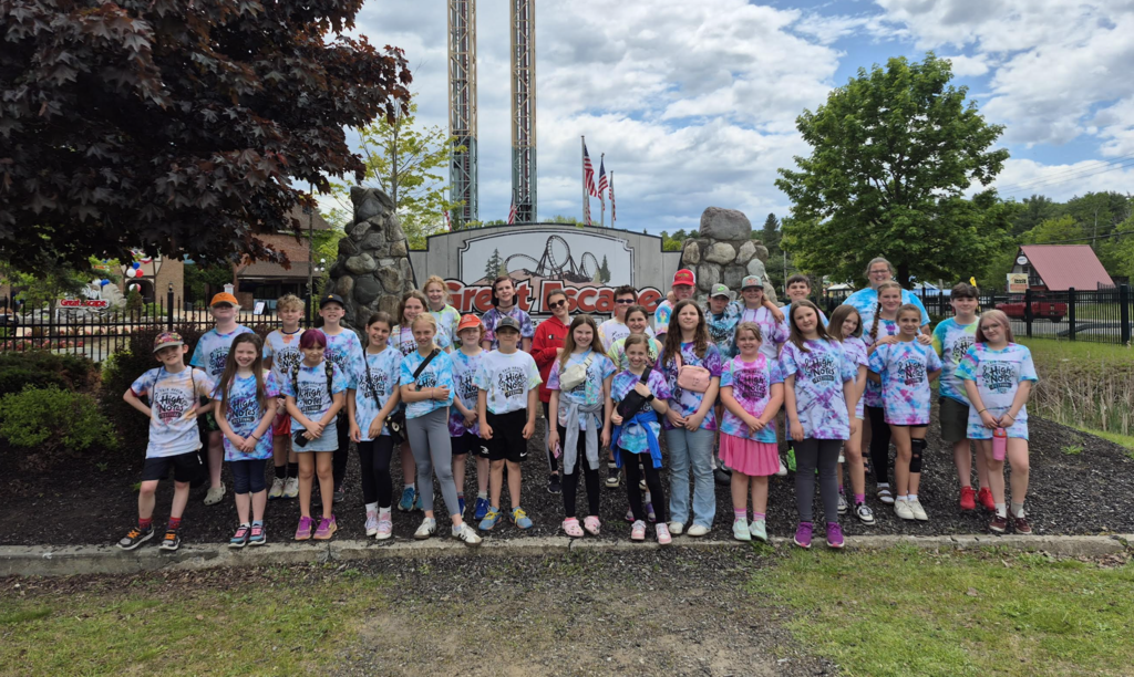 A large group of Fair Haven Grade School students and chaperones wearing matching 2026 High Notes Festival tie-dye shirts pose for a group photo in front of the Great Escape amusement park entrance on a cloudy day. Roller coaster structures and park signage are visible in the background.