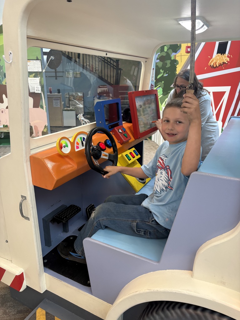 A young student sits in the driver's seat of a large interactive vehicle exhibit, holding a rope control while operating buttons, screens, and a steering wheel. An adult stands behind him.