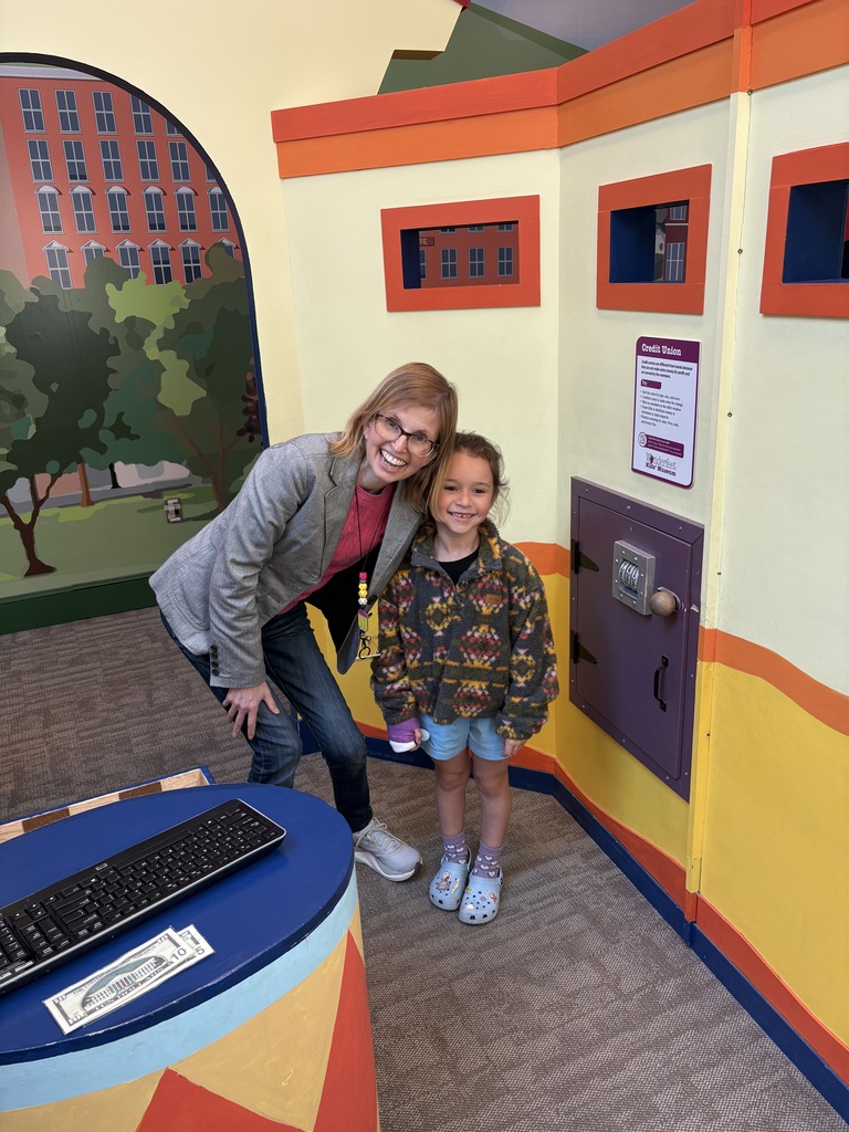 An adult and a smiling young student pose together inside a colorful pretend credit union exhibit at the Wonderfeet Kids Museum. A play computer keyboard and paper money sit on a nearby counter.