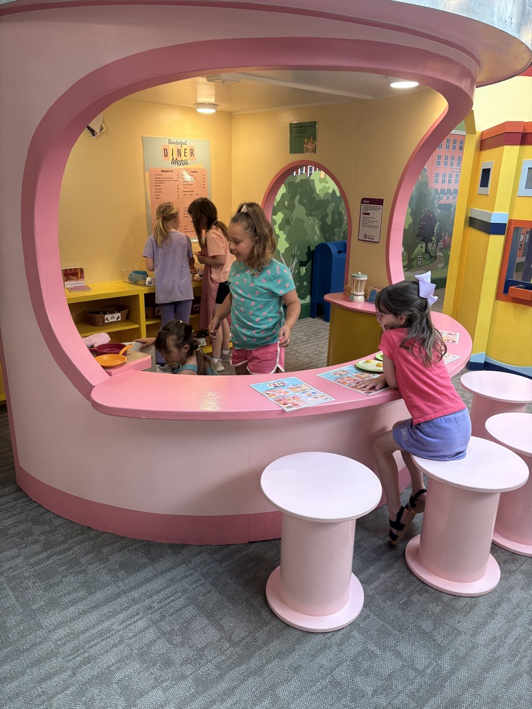 Several students play together inside a brightly colored diner exhibit, pretending to prepare and serve food while sitting and standing around a pink counter.