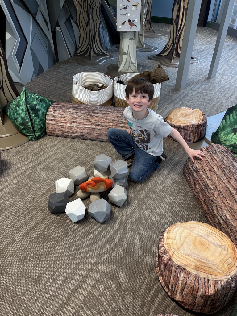 A smiling student kneels beside a pretend campfire made of soft rocks and logs in a woodland-themed exhibit area of the museum.