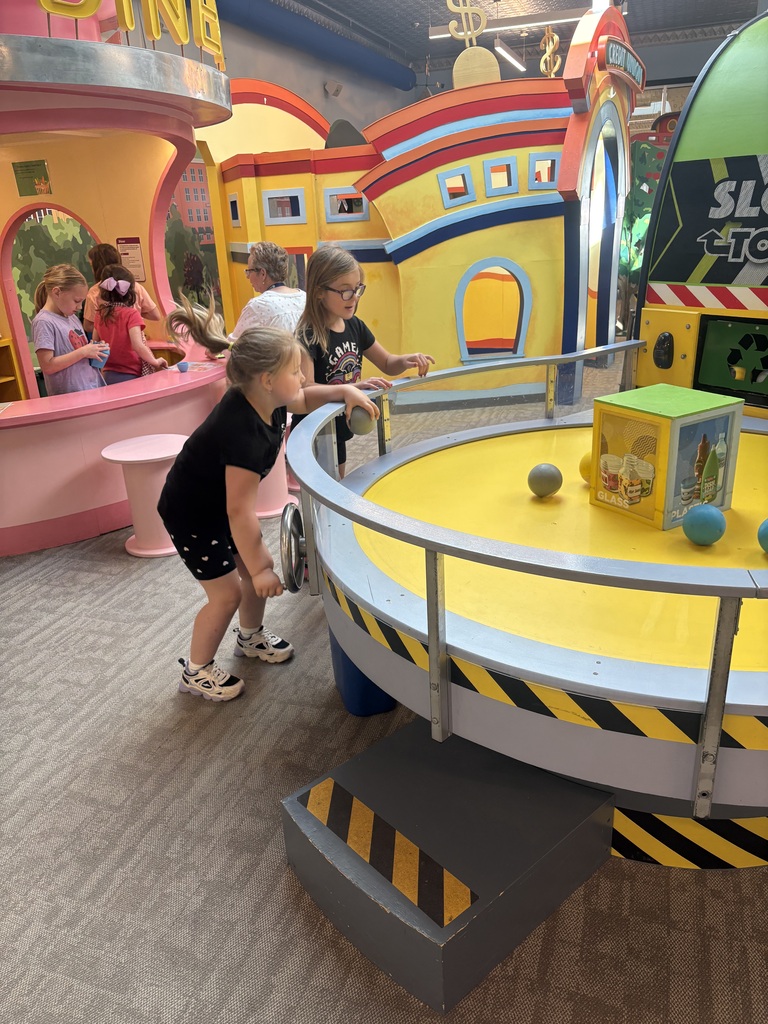 Students interact with a recycling-themed exhibit, rolling balls around a circular table while learning about sorting materials. Other children play in nearby exhibits.