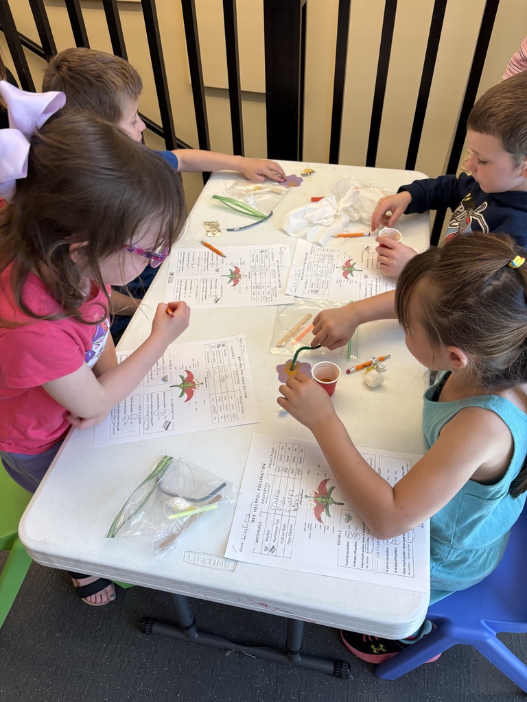 Several first-grade students sit around a table during a pollination activity, examining materials and completing worksheets while building hand pollinators.