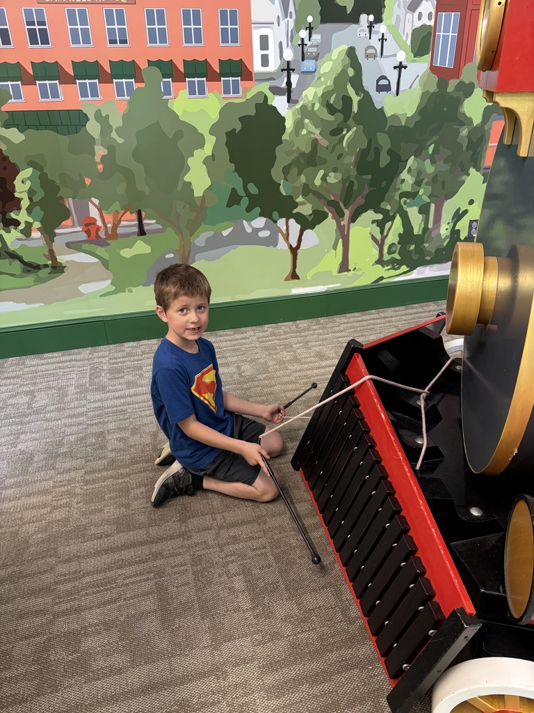 A student sits beside a large train-themed musical exhibit, holding mallets and preparing to strike the instrument.