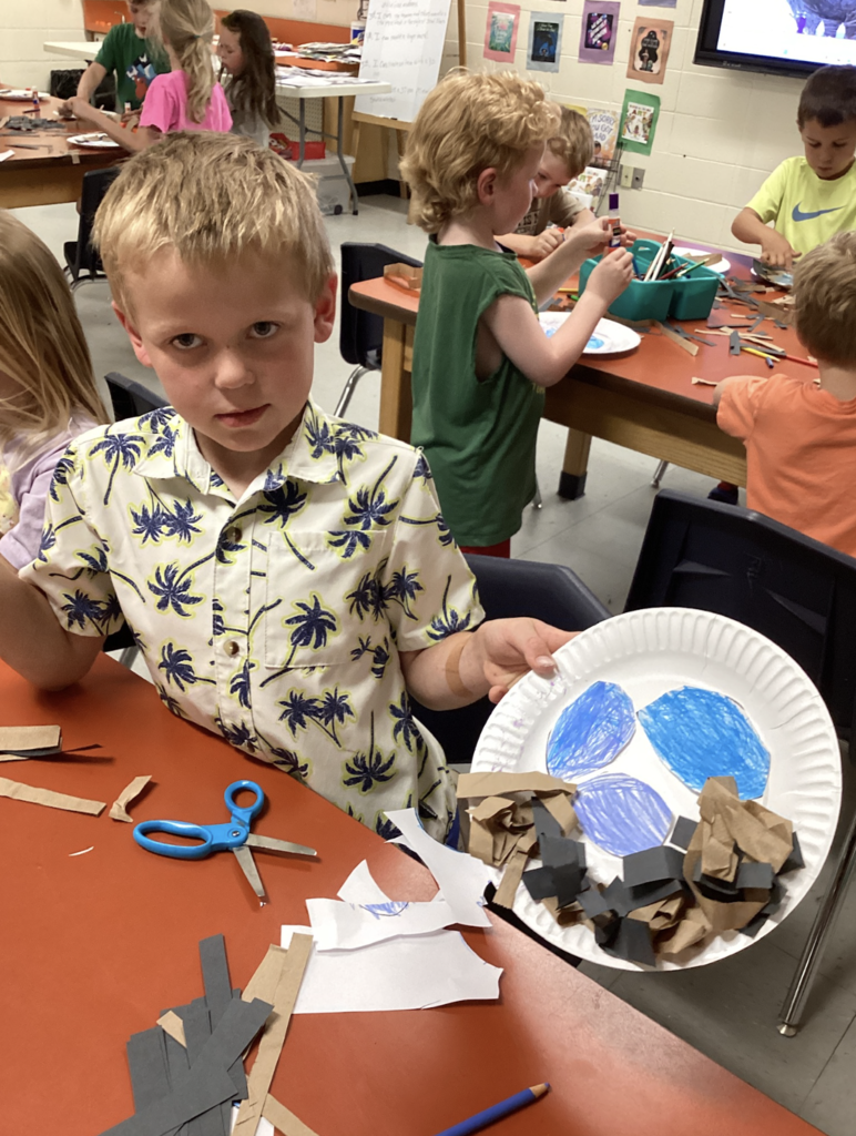 A kindergarten student wearing a patterned shirt proudly displays his completed bird nest craft featuring blue eggs and crumpled paper materials. Other students work on similar projects in the background.
