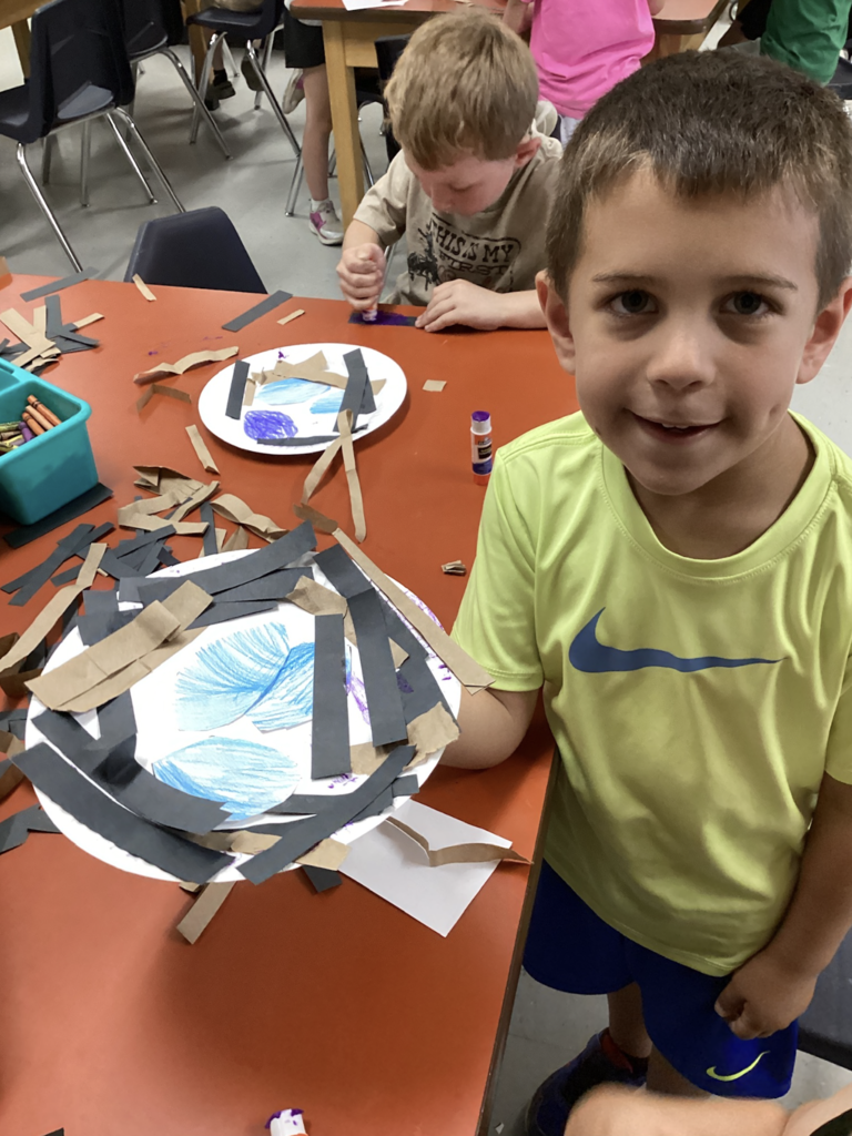 A smiling kindergarten student holds up his completed bird nest craft made from a paper plate, blue eggs, and layered paper strips. Another student in the background continues decorating a similar project.