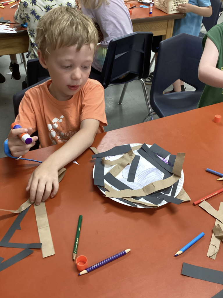 A kindergarten student concentrates on gluing strips of brown and black paper onto a paper plate nest while seated at a classroom table covered with art supplies and craft materials.