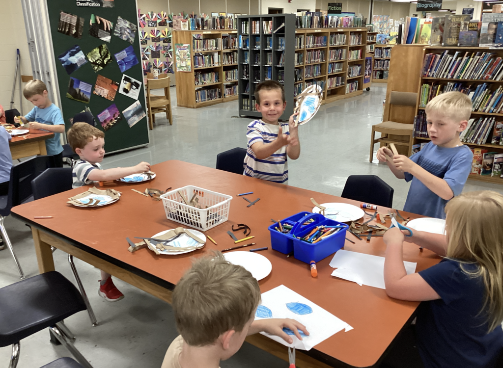 Kindergarten students work on bird nest crafts at a large table in the school library. One student proudly holds up his paper plate nest while classmates cut, glue, and decorate their own creations among bookshelves and library displays.