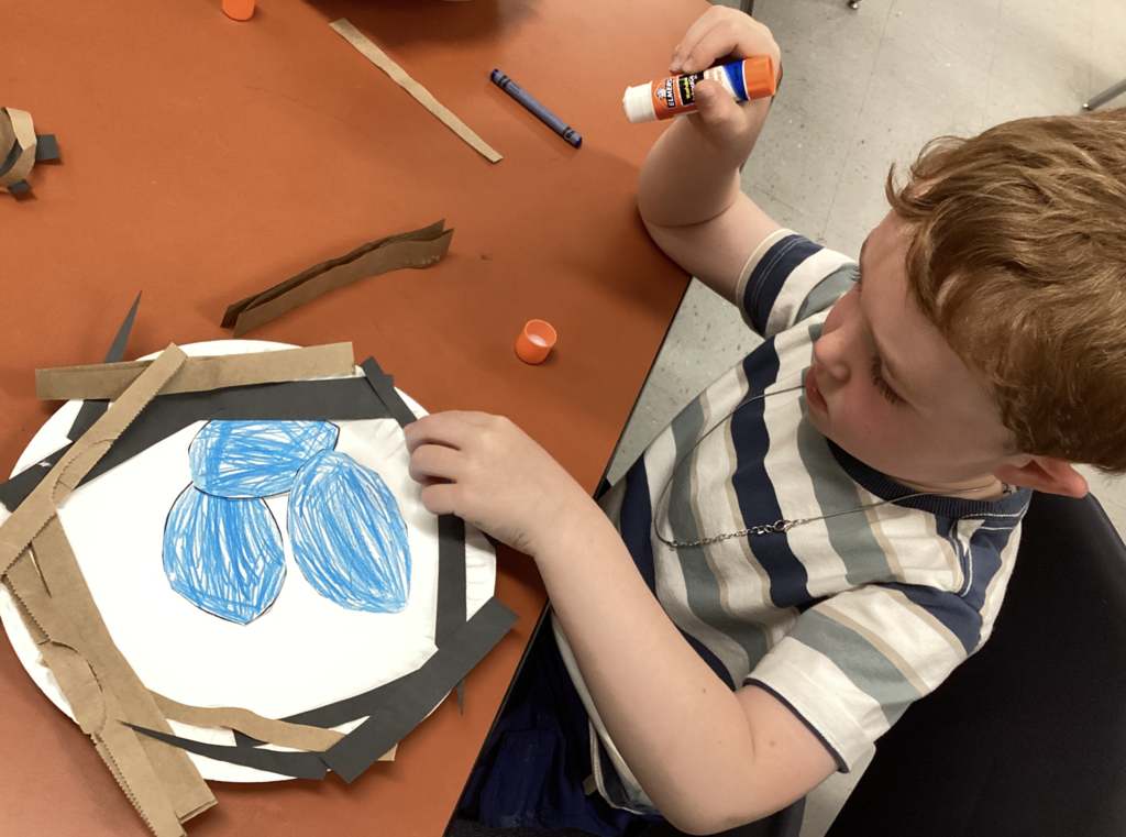 A close-up of a kindergarten student carefully gluing paper strips around a paper plate nest. The nest includes blue crayon-colored eggs in the center and layered paper pieces around the edge.