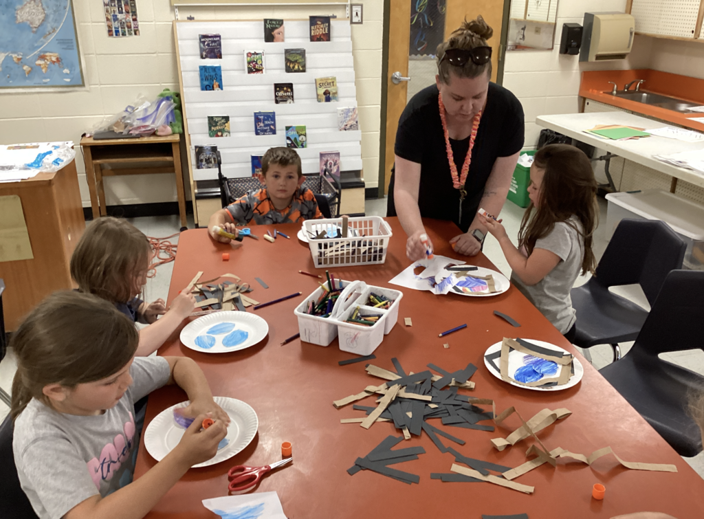 A teacher assists kindergarten students as they build paper plate bird nests. Students use glue, crayons, and paper strips at a table in the library classroom while working on the hands-on activity.