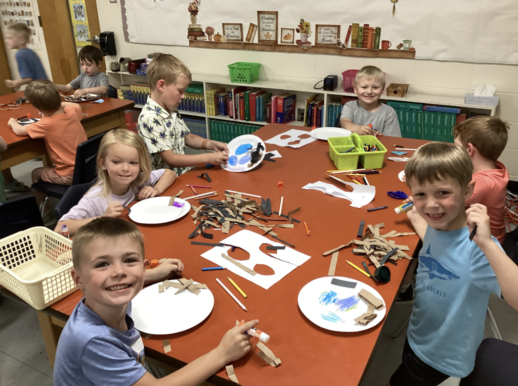 Kindergarten students sit around a classroom table covered with craft supplies, paper plates, crayons, glue sticks, and strips of paper while creating bird nests inspired by The Nest That Robin Built. Several students smile at the camera while working on their projects.