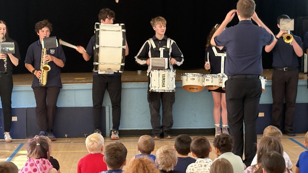 A student conductor leads members of the Fair Haven Union Middle and High School band during a performance for elementary students. Band members play percussion, woodwind, and brass instruments while younger students watch attentively from the gym floor.