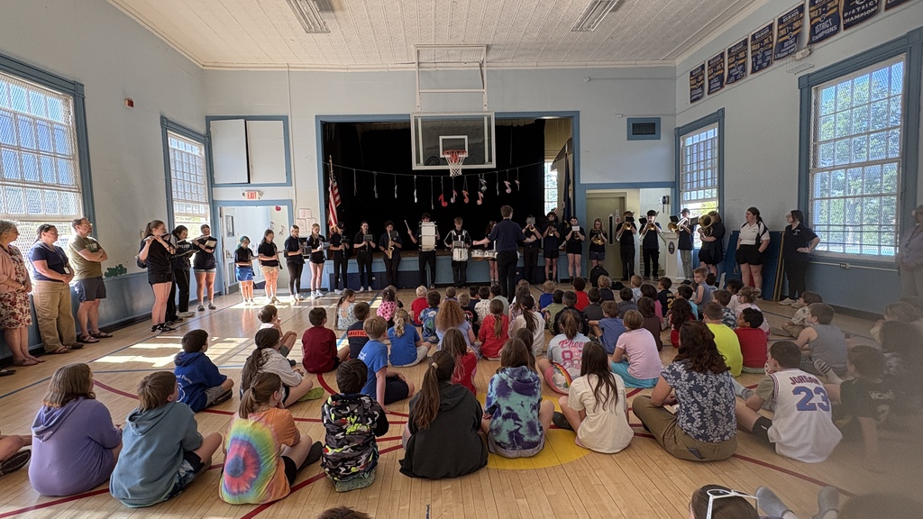 Wide view of a school gymnasium as the Fair Haven Union Middle and High School band performs for a large group of elementary students seated on the floor. Teachers and staff stand along the walls while student musicians play a variety of instruments at the front of the room beneath hanging music note decorations.