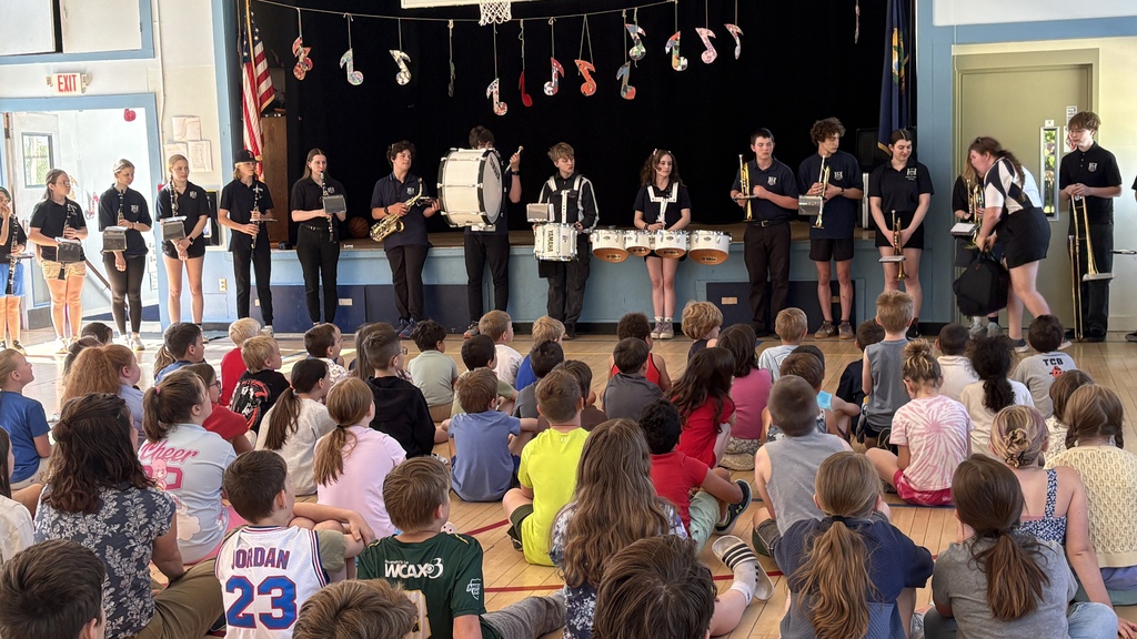 Fair Haven Union Middle and High School band students stand at the front of a school gymnasium holding a variety of instruments, including clarinets, saxophones, drums, trumpets, and trombones, while elementary students sit on the floor watching. Colorful music note decorations hang above the stage area during the performance.