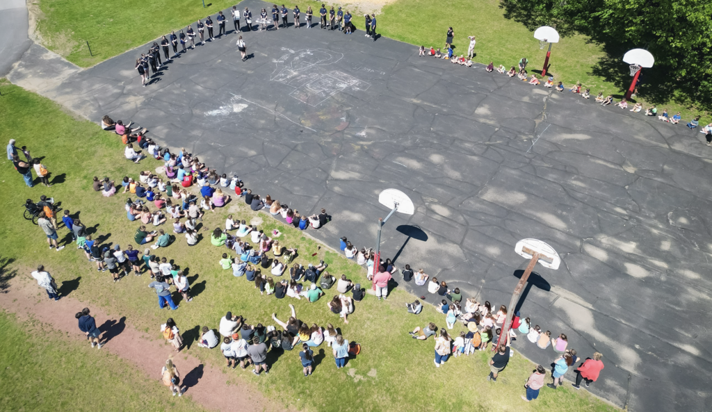 Aerial view of Castleton Elementary School students and staff gathered around a large playground area while FHUMHS band students perform and demonstrate instruments from the center. Basketball hoops and groups of seated students are visible around the perimeter of the blacktop.