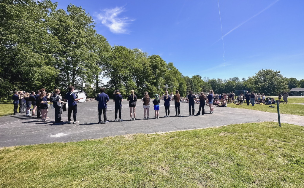 Wide outdoor view of FHUMHS band students arranged in a large semicircle on a paved playground while elementary students and staff sit and stand nearby listening. The presentation takes place on a sunny day beneath a bright blue sky.