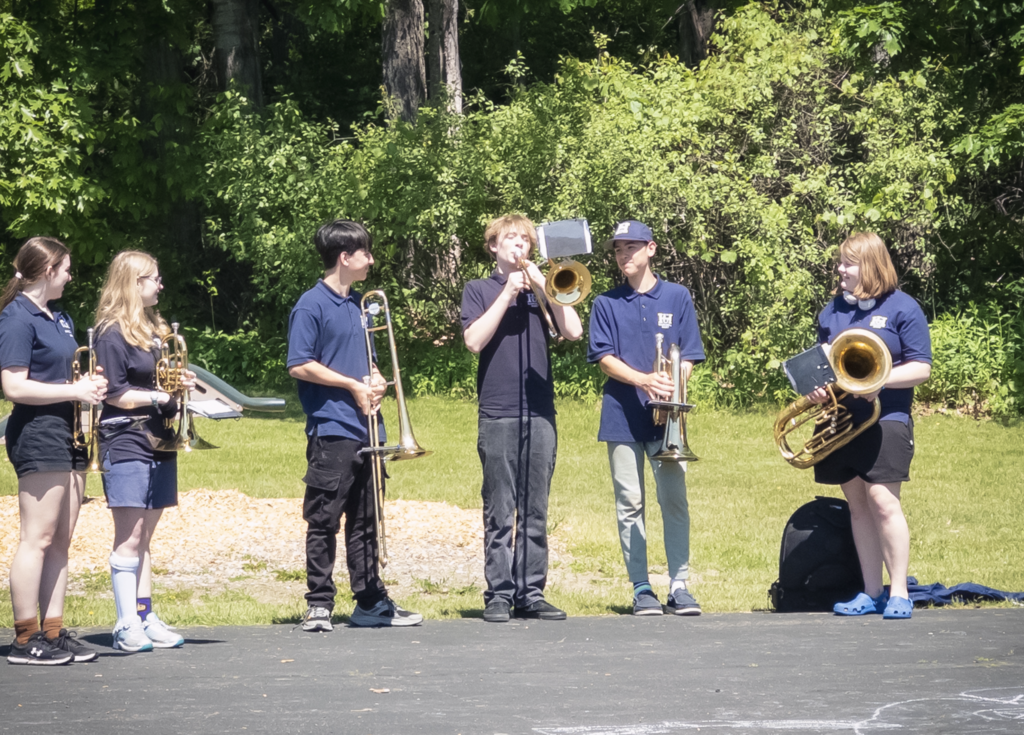 A group of FHUMHS band students stand outdoors holding trumpets, trombones, and a tuba. One student plays a trombone while the others listen and prepare to demonstrate their instruments.